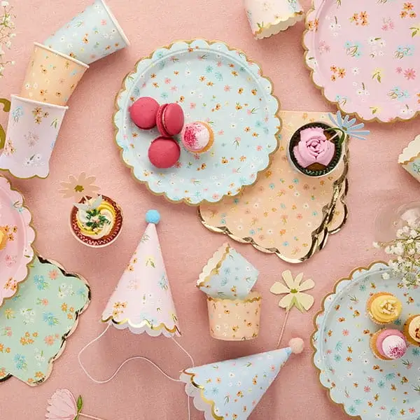 Floral tea party tableware with pastel plates, cups, party hats, and small cakes arranged on a pink background